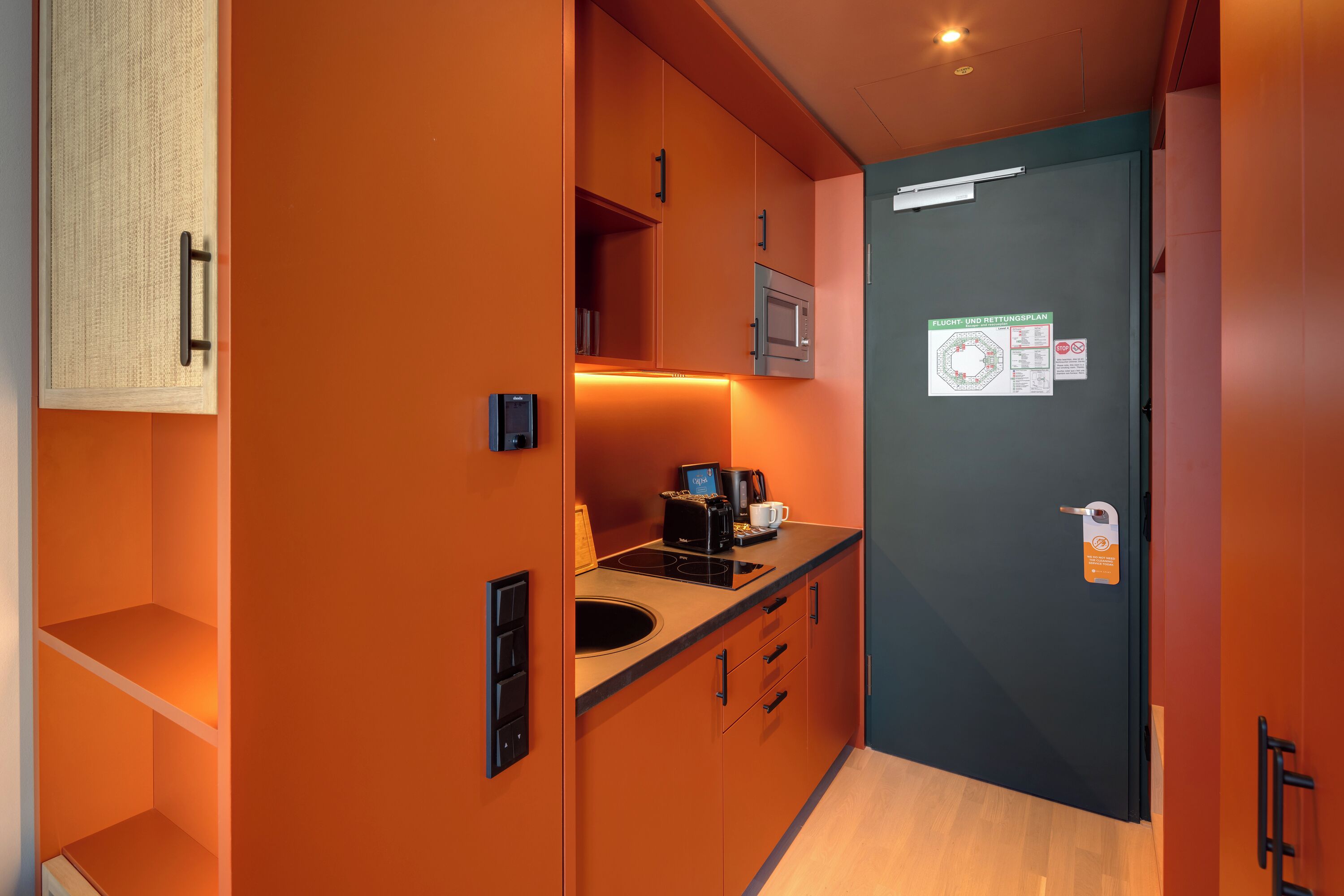 Une cuisine compacte équipée d’un four à micro-ondes, d’une machine à café et d’un petit évier. Une porte avec une pancarte et un interrupteur sont fixés au mur.