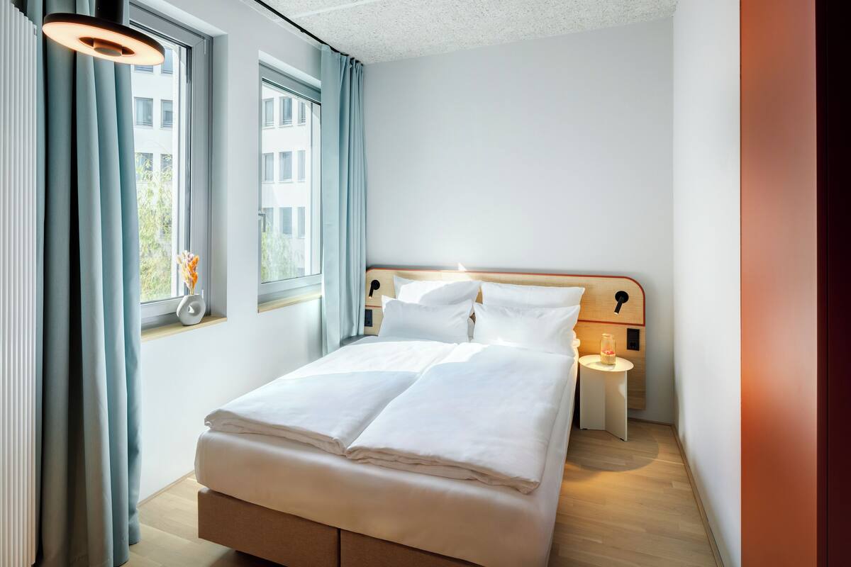A neatly made bed in a hotel room with a wooden headboard, a bedside table with a lamp, and a window with curtains.