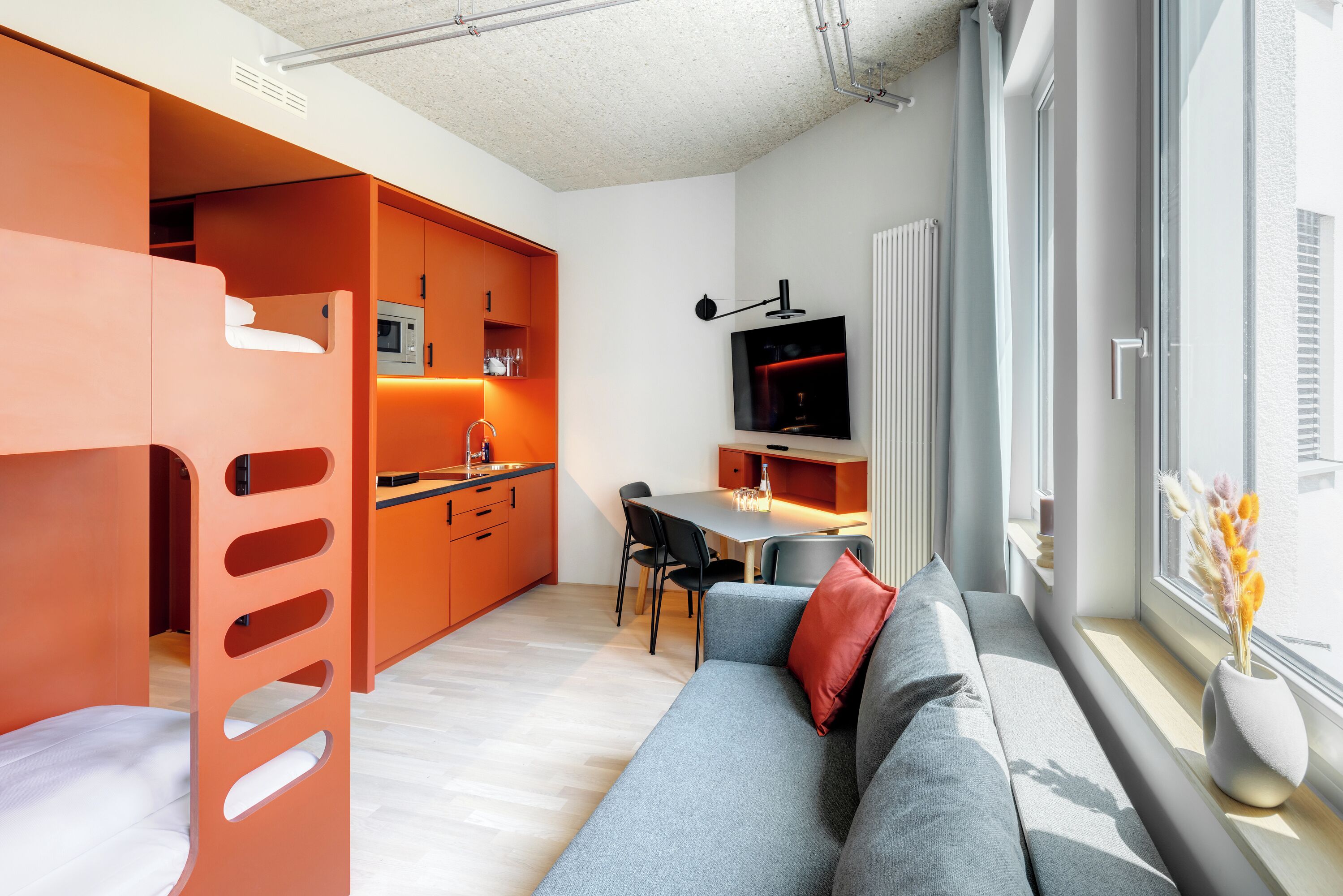 Un espacio habitable moderno con un sofá gris, cocina naranja y una zona de comedor con mesa y sillas.