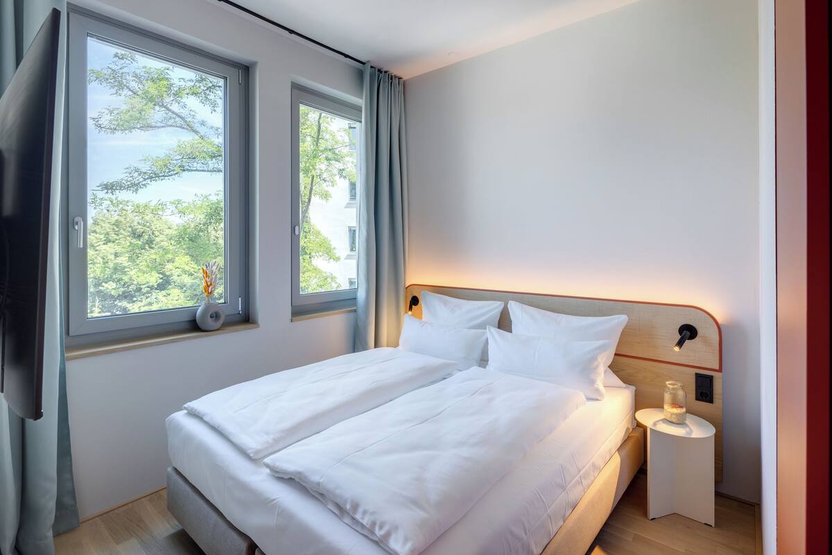 A neatly made bed with white linens, a bedside table with a lamp, and a window with curtains.