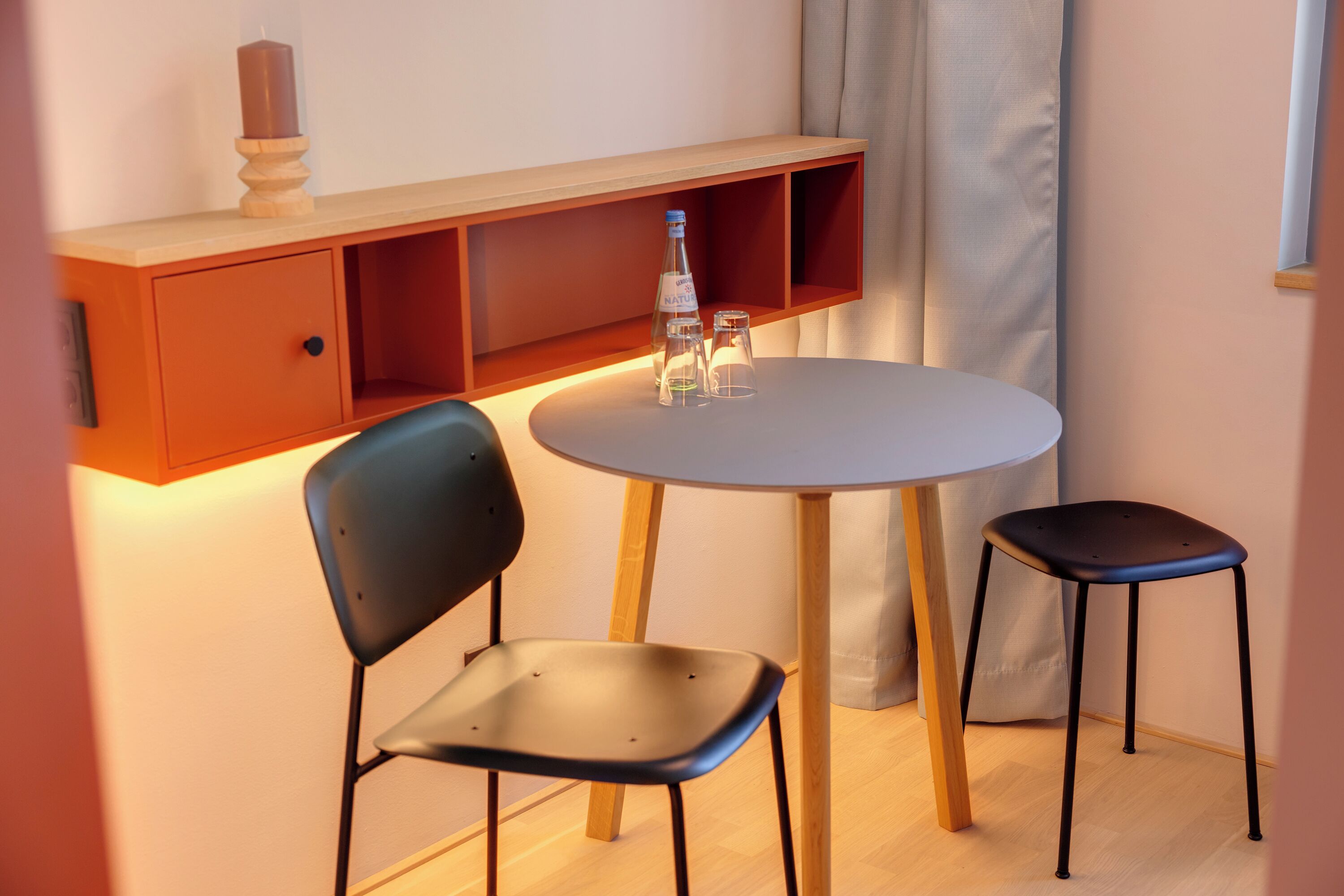 Une petite pièce avec une table ronde, deux chaises, une étagère avec une bougie et une bouteille.