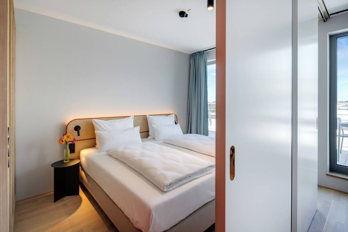 A modern hotel room with a large bed, a bedside table with a vase of flowers, and a view of the city through the window.