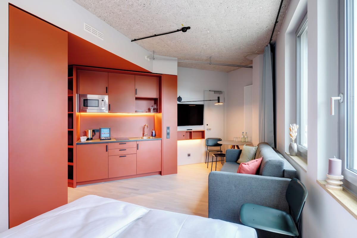 A modern, compact living space with a kitchenette, a sofa, a dining area, and a large window providing natural light.