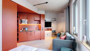 A modern, compact living space with a kitchenette, a sofa, a dining area, and a large window providing natural light.