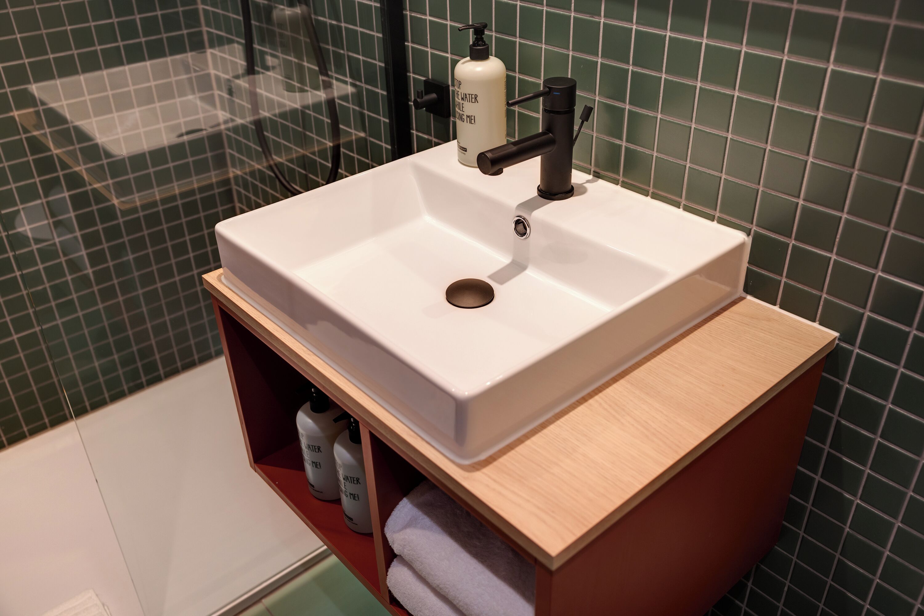 Een badkamer met een wit wastafel, een houten wastafelmeubel en een muur betegeld met groene tegels.