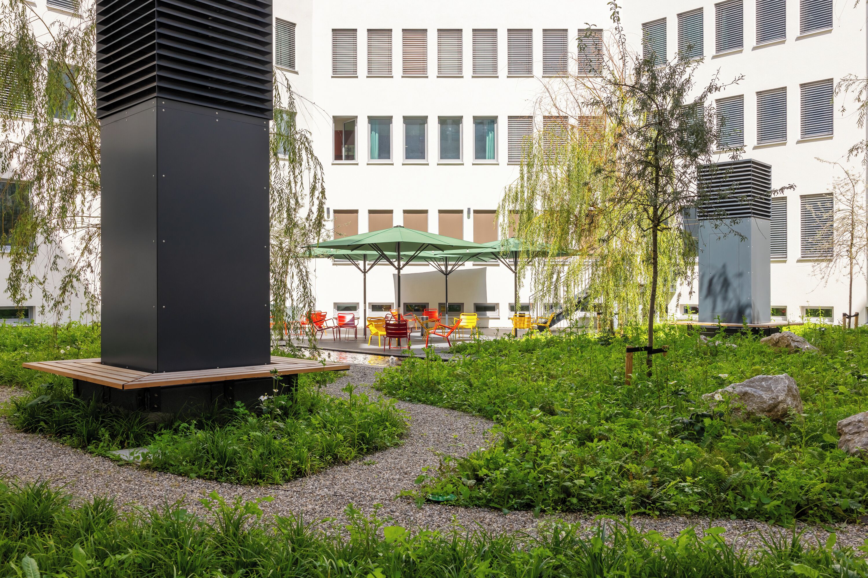 A courtyard with a black structure, green umbrella seating, and red and yellow chairs.