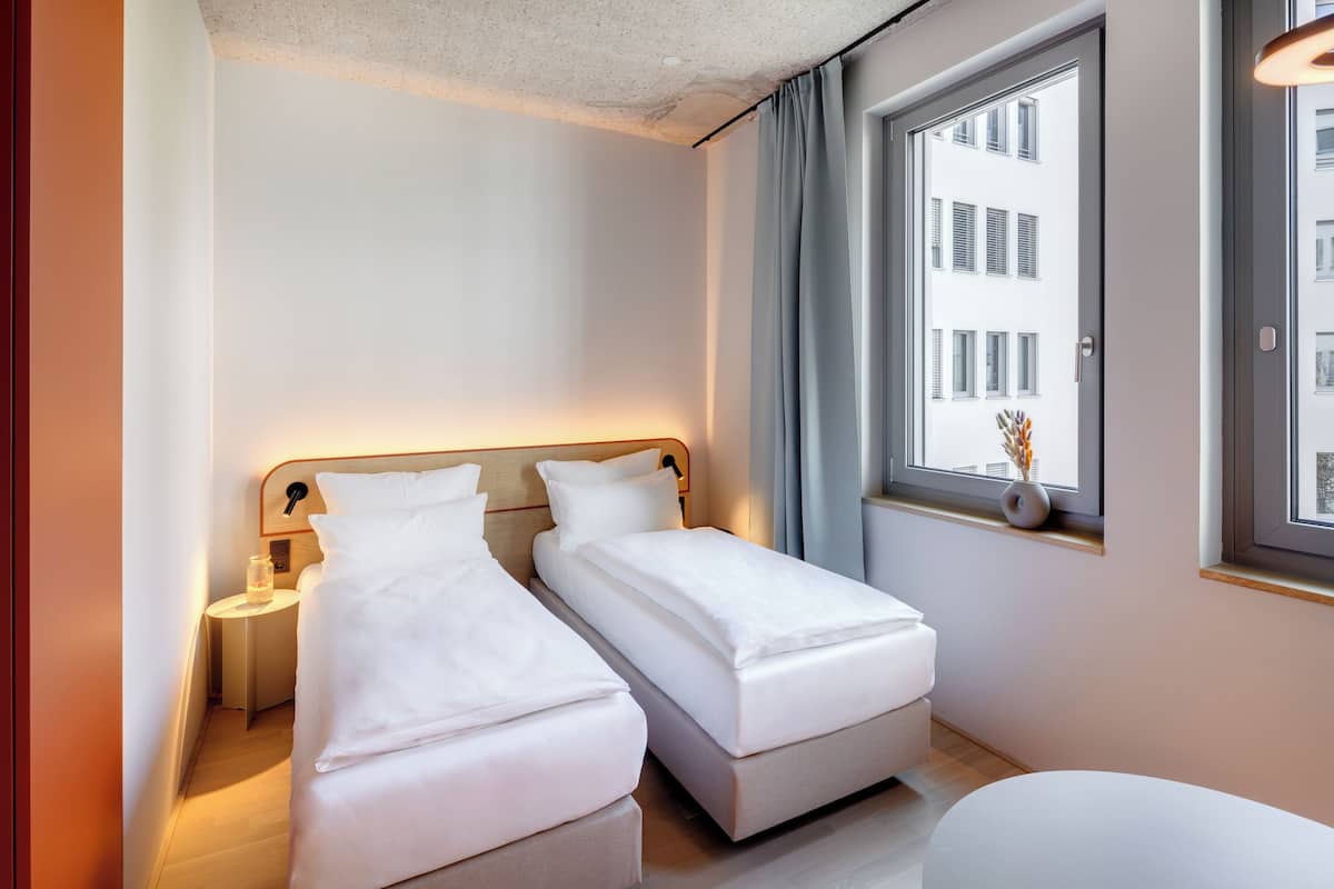 A modern hotel room with two single beds, a bedside table with a lamp, a window with a view of buildings, and a small table with a vase.
