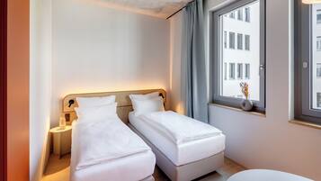 Habitación de hotel moderna con dos camas individuales, una mesita de noche con lámpara, una ventana con vistas a edificios y una pequeña mesa con un florero.
