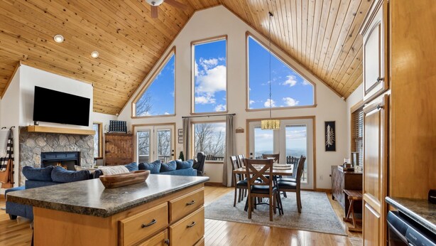 A spacious living area with a large wooden ceiling, a stone fireplace, a flat-screen TV, a kitchen with wooden cabinets, and a dining area with a table and chairs. - Private Mountain Cabin Retreat 1.5 acres, Amazing Views, Hot Tub, STARLINK Wifi (Murphy)
