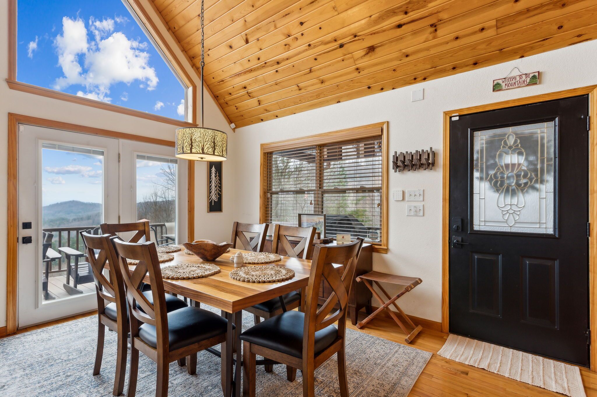 Un comedor con una mesa y sillas de madera, una puerta negra con un panel de vidrio decorativo y vistas a las montañas a través de amplios ventanales.