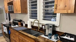 Una cocina con armarios de madera, encimera negra, un lavabo, cafetera y una ventana con persianas.