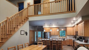 Une cuisine spacieuse avec des meubles de rangement en bois, un coin repas avec une longue table et des chaises en bois, et un escalier en bois menant à l’étage supérieur.