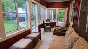 Une véranda meublée de mobilier en osier, dotée de grandes fenêtres et offrant une vue sur un jardin verdoyant.