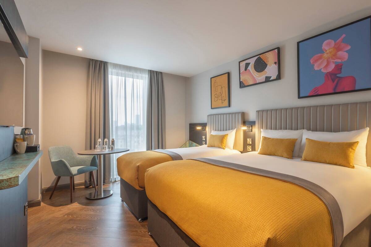 Une chambre d’hôtel avec un grand lit, deux tables de chevet, un petit bureau avec une chaise et une fenêtre avec des rideaux.