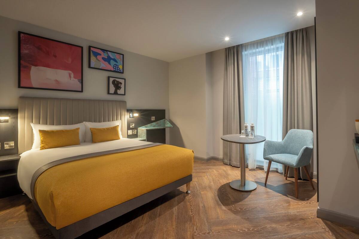 Une chambre d’hôtel avec un lit, une petite table ronde, une chaise et une vue sur la ville grâce à de grandes fenêtres.