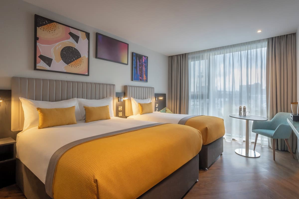 A hotel room with two beds, a dining area, and large windows with curtains.