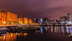 A nighttime cityscape with buildings reflecting on calm water, a bridge, and a river.