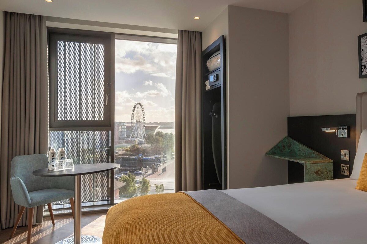 A hotel room with a bed, a table, a chair, a TV, and a view of a Ferris wheel.