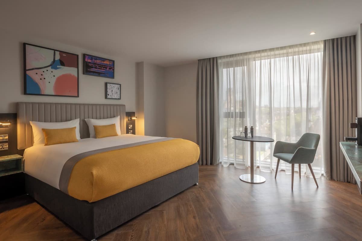 A modern hotel room with a large bed, a small round table, two chairs, a desk with a computer, and a large window with curtains.