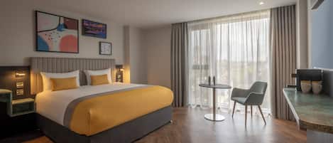 A modern hotel room with a large bed, a small round table, two chairs, a desk with a computer, and a large window with curtains.
