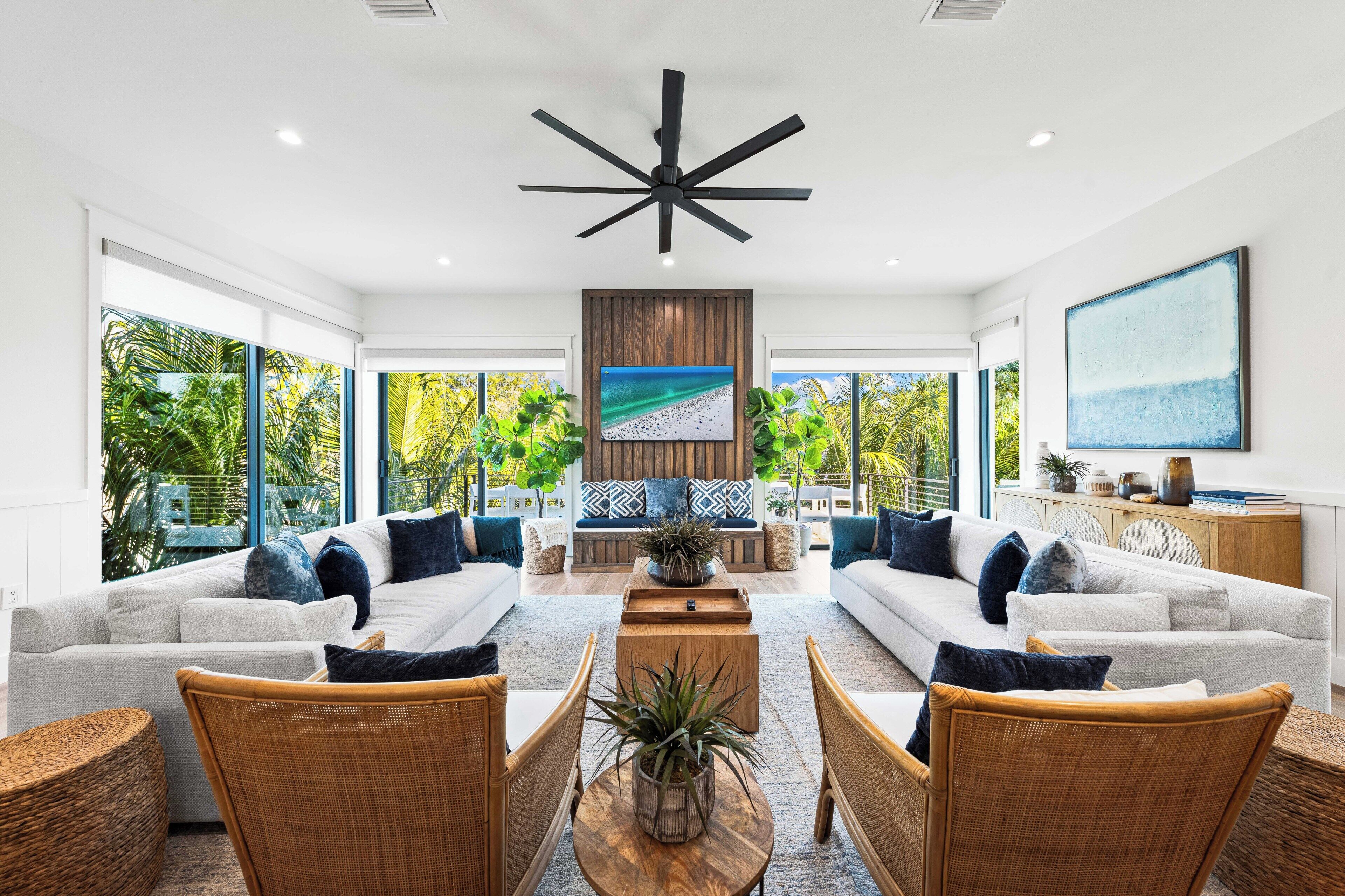 Amplia sala de estar con un ventilador de techo grande, sofás blancos con cojines azules, sillas de mimbre y una mesa de centro de madera.
