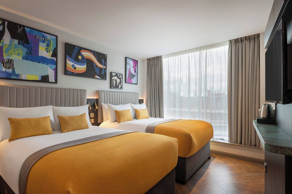 A hotel room with two beds, a large window with curtains, and artwork on the walls.