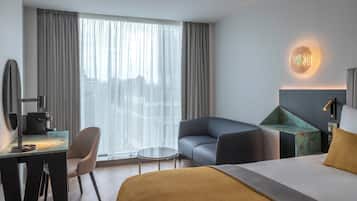 Habitación de hotel con cama, escritorio con cafetera, silla, sofá y ventana grande con cortinas.