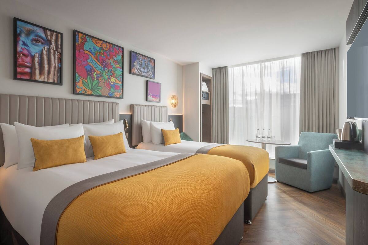 A hotel room with two beds, a desk, a chair, and artwork on the walls.
