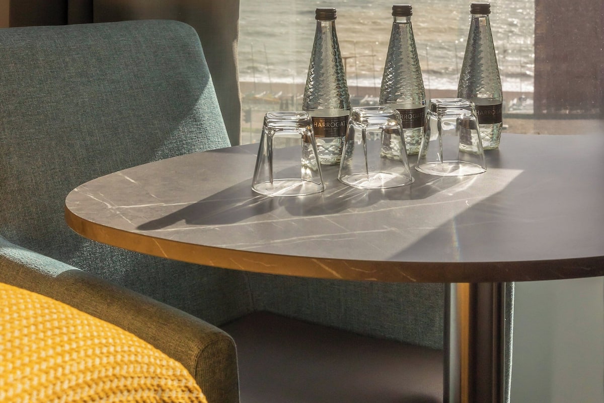 A room with a view of the ocean, a table with glasses and bottles, and a sofa.