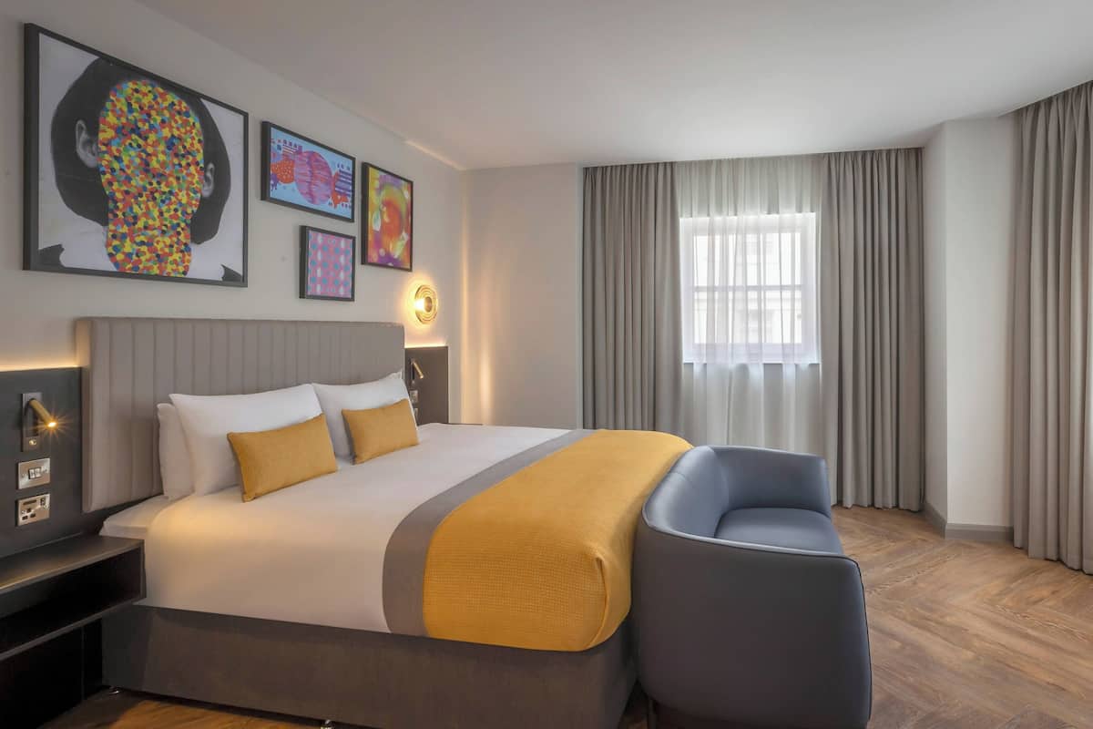 A modern hotel room with a large bed, a bedside table with a lamp, a chair, and a window with curtains.