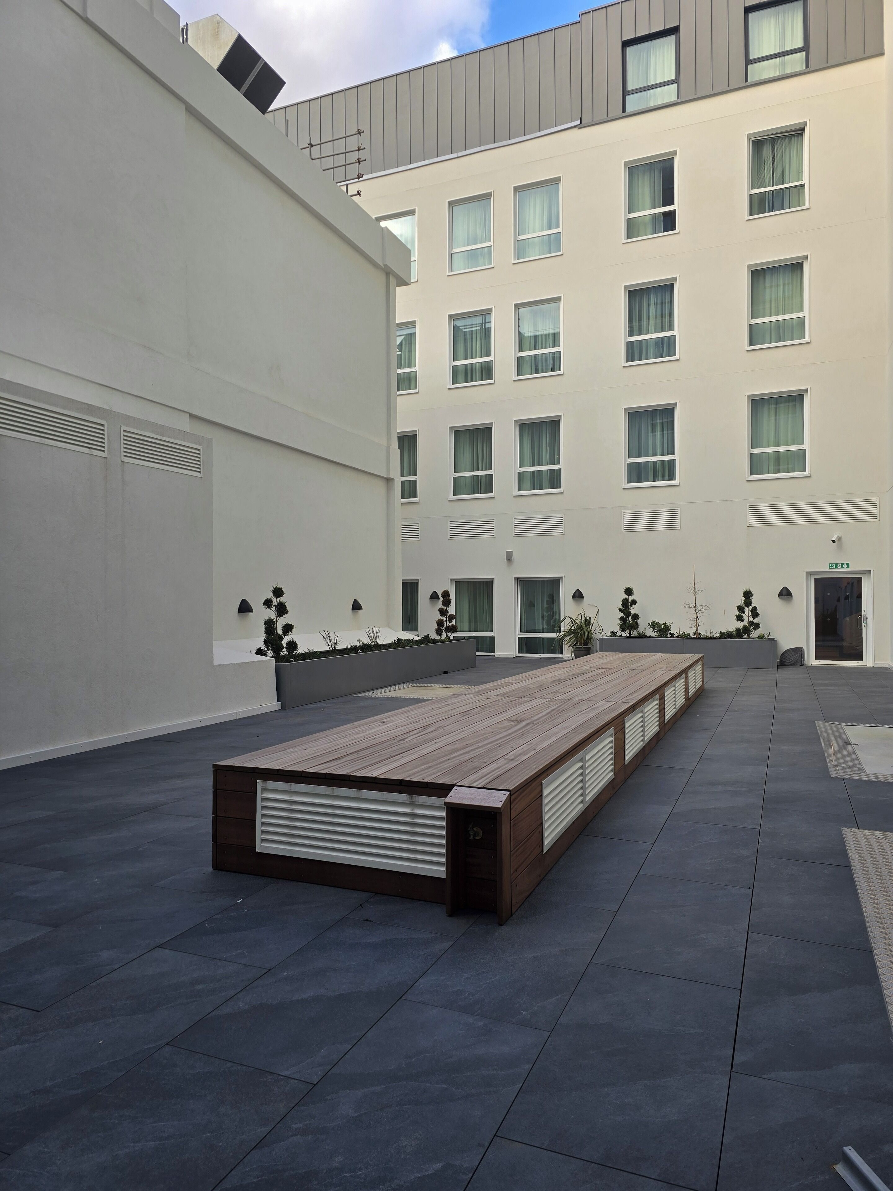 A modern building with a flat roof, multiple windows, and a long wooden bench in the courtyard.