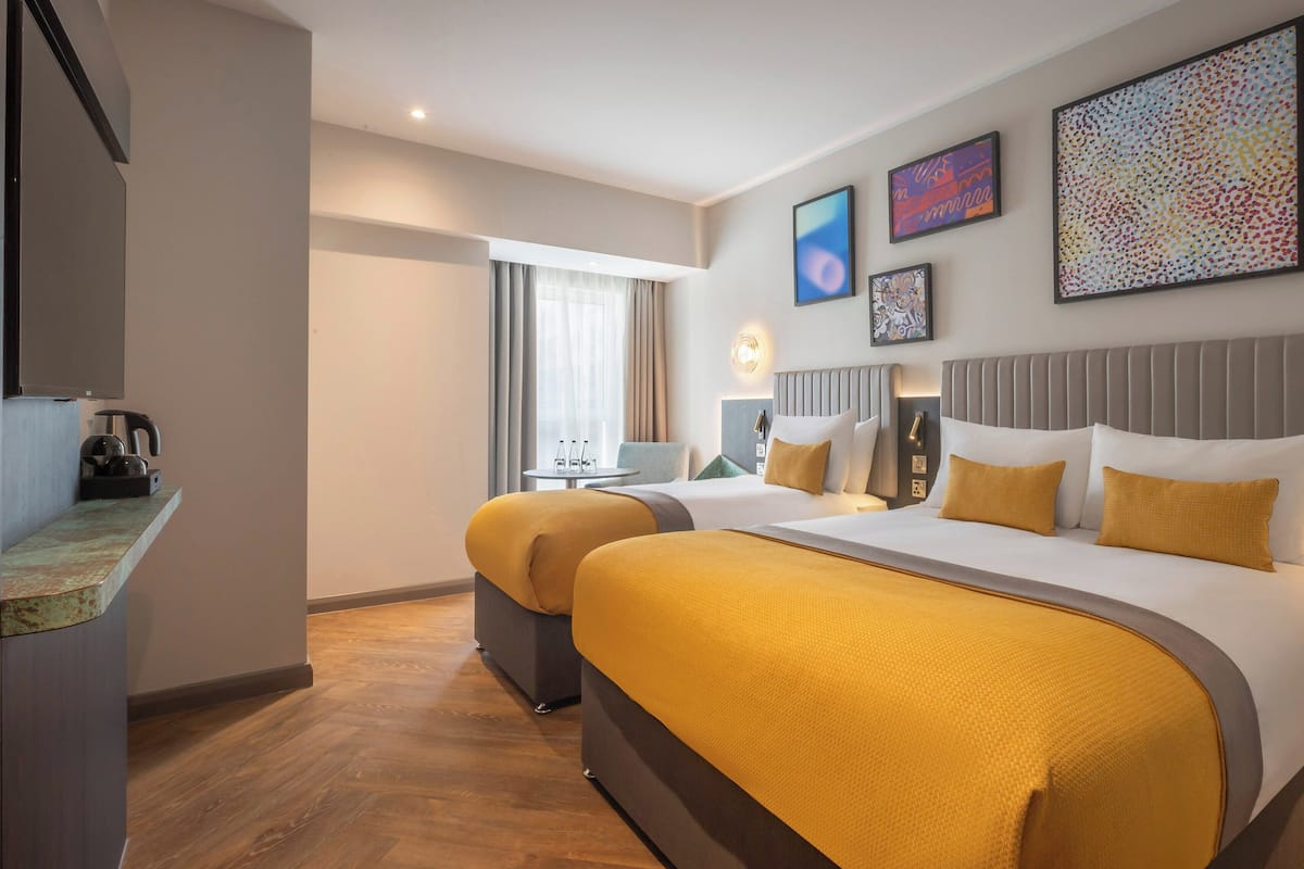 A hotel room with a large bed, two bedside tables, a desk, and a chair. There are two windows with curtains, a wall-mounted TV, and artwork on the wall.