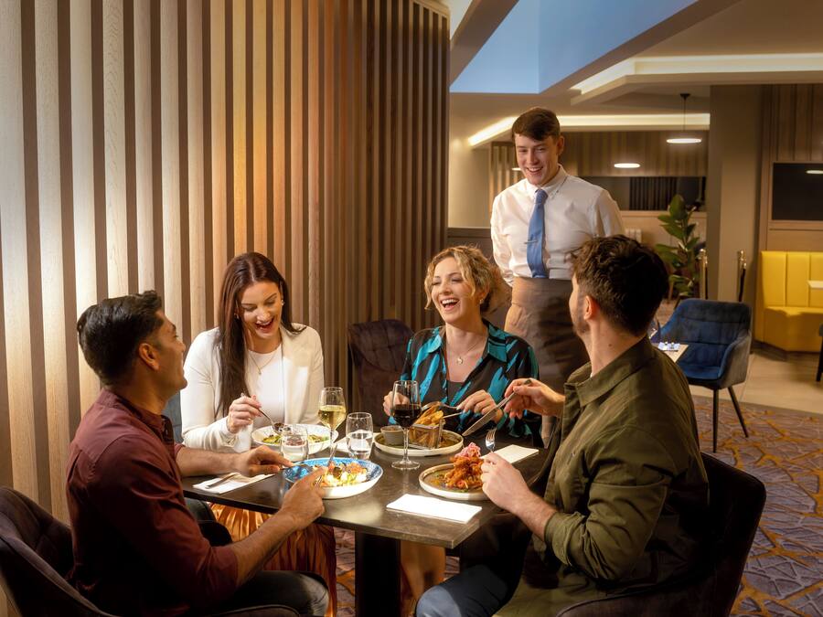 A group of people dining at a table in a modern hotel room with a striped wall and a patterned carpet.