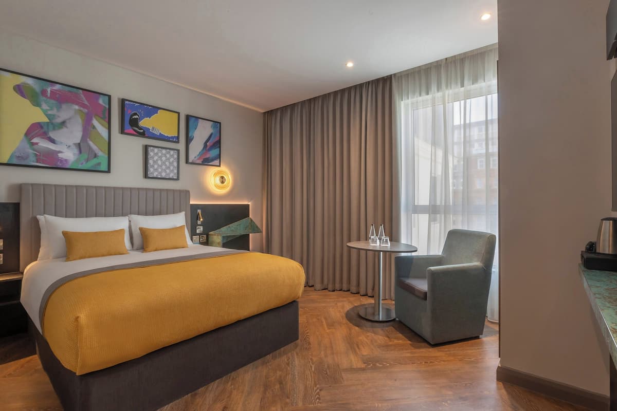 A hotel room with a bed, a chair, a small table, and artwork on the wall.