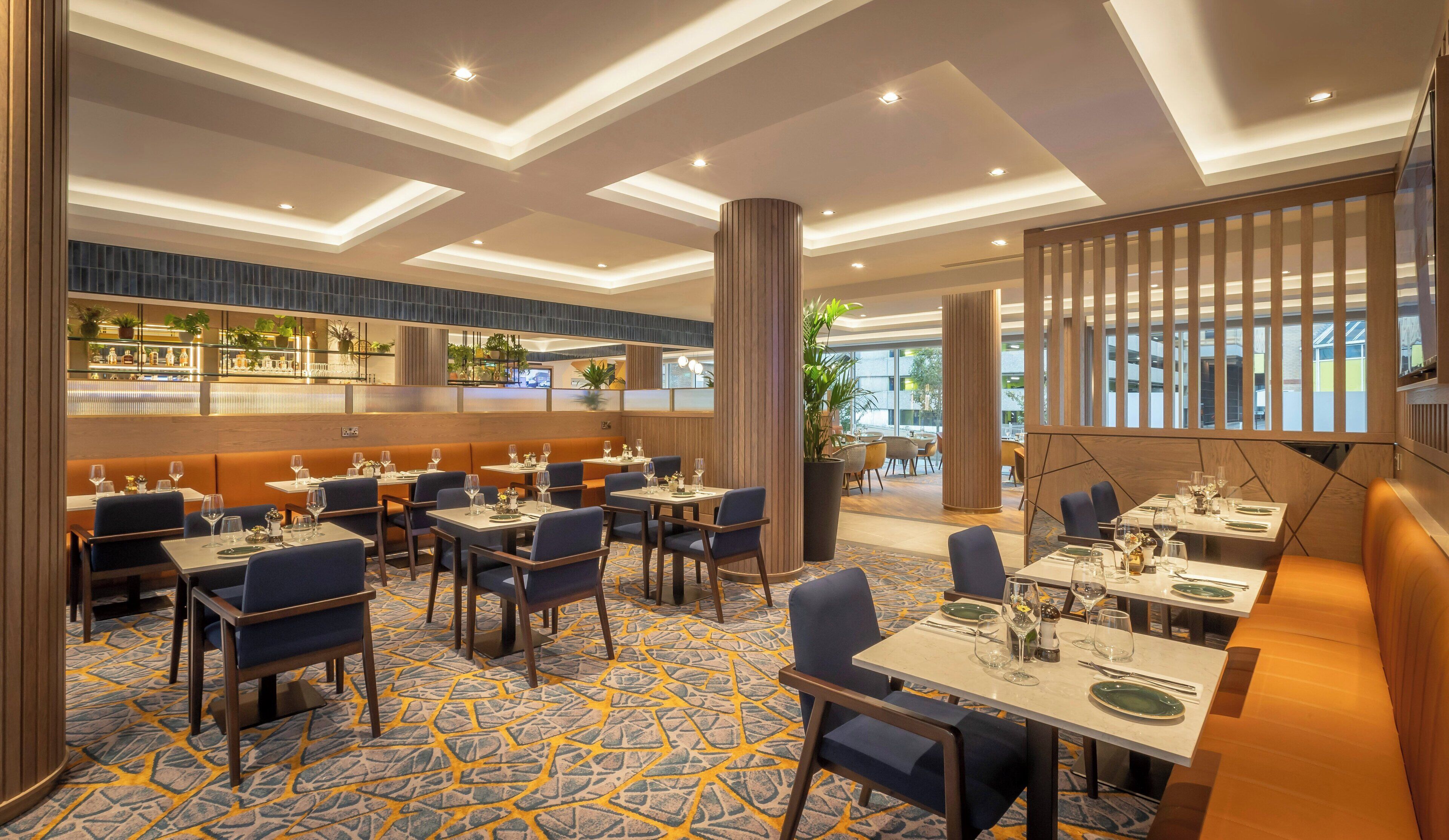 A modern restaurant with a central bar area, multiple dining tables, and a patterned carpet.
