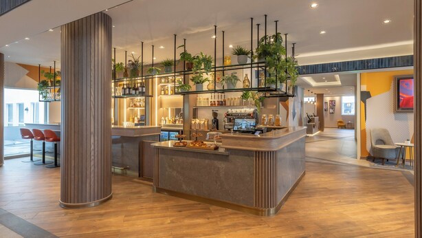 A modern bar area with a wooden floor, a central bar counter, and a variety of plants and bottles on shelves. - Maldron Hotel Brighton City Centre (Brighton)
