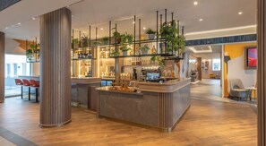 A modern bar area with a wooden floor, a central bar counter, and a variety of plants and bottles on shelves. - Maldron Hotel Brighton City Centre (Brighton)