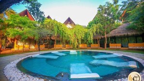 A circular pool surrounded by a pebble border, with thatched-roof structures and lush greenery in the background.