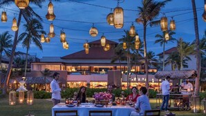 An outdoor dining area with string lights, palm trees, and a building in the background.