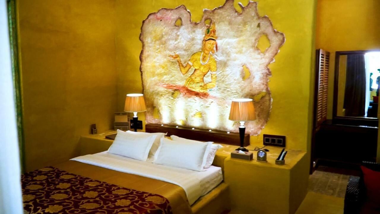 Habitación de hotel con una cama, dos lámparas de noche, una mesita de noche con un reloj y un elemento decorativo grande en la pared.