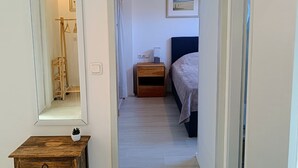 Een gang die leidt naar een slaapkamer met een bed, nachtkastje, spiegel en een klein houten kastje.