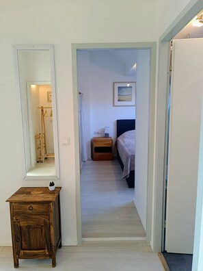 Un couloir mène à une chambre qui contient un lit, une table de chevet, une commode, un miroir et un petit meuble en bois.