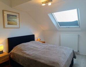 Une chambre à coucher avec un lit, une table de chevet avec une lampe, un tableau encadré au mur, un puits de lumière et un radiateur.