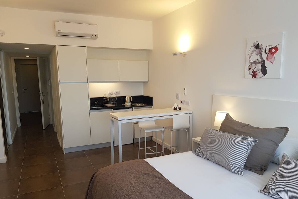 Ein kleines Hotelzimmer mit Küchenzeile, einem Bett mit gestreiften Kissen und einem Tisch mit Stühlen.
