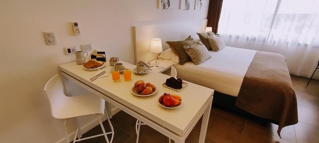 A hotel room with a bed, a bedside table with breakfast items, a chair, and a lamp.