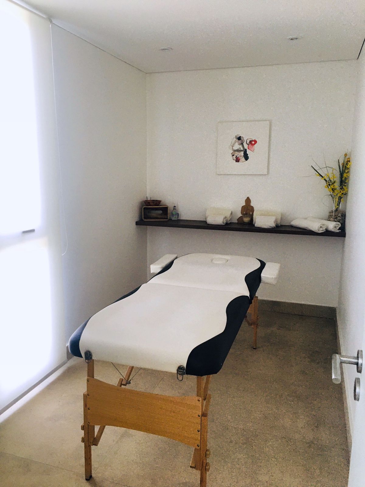 A treatment room with a wooden table, a shelf with towels and a Buddha statue, and a framed artwork on the wall.