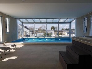Piscina interior com paredes de vidro transparente, cadeiras de descanso e vista para os edifícios ao redor.