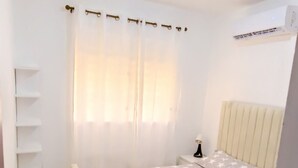 A bedroom with a bed, a bedside table, a nightstand, a window with curtains, a radiator, and an air conditioning unit. - Apto, Res, Carmen Renata Iii, a 8 Minutos de la Embajada Americana sto dgo (Pantoja)