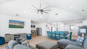 Un'ampia zona giorno con una moderna ventola a soffitto, una TV a schermo piatto montata a parete, una sala da pranzo con sedie blu e una cucina con pensili bianchi.