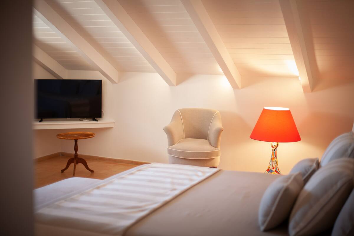 A bedroom with a bed, a chair, a table, a lamp, and a TV.